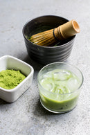 Bowl with matcha tea whisk, matcha powder, and chilled matcha drink in a glass.